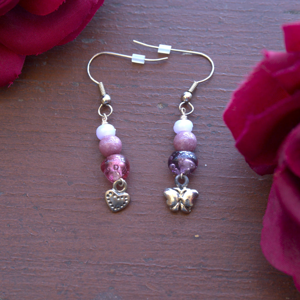Pair of beaded earrings with a butterfly charm on one and a heart charm on the other on a wooden surface with red flowers.