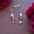 Pair of beaded earrings with a butterfly charm on one and a heart charm on the other on a wooden surface with red flowers.
